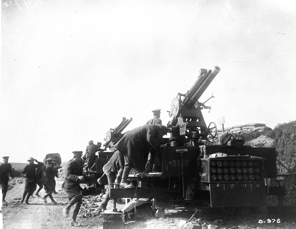 This photograph shows a 13 pounder 9 cwt anti-aircraft gun, on a mobile mounting, with a rack of ammunition in the foreground. Crews appear to be scrambling to ready the guns. MIKAN No. 3395208